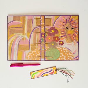 Carnet SUZANA posé ouvert avec un stylo rose et un marque-page en bois à côté.